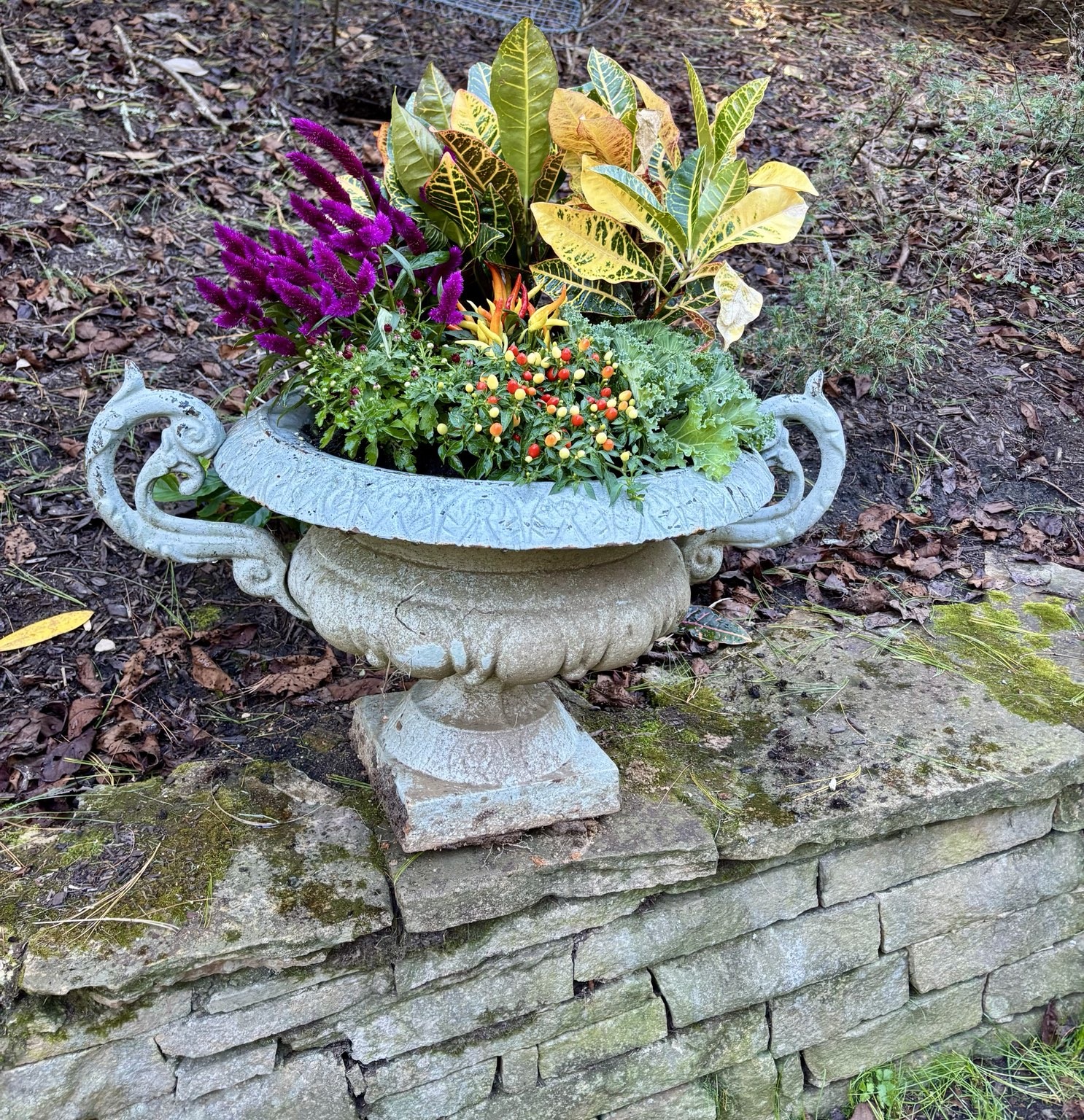 Ornate pale stone bowl filled with fall flowers beside a stone wall