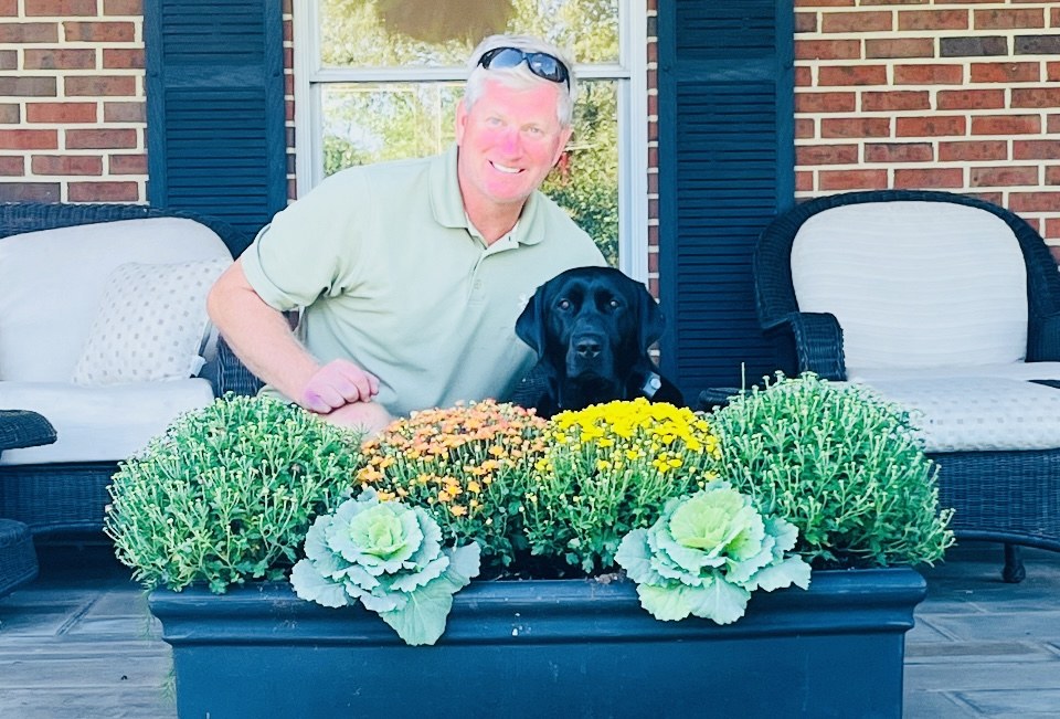 Derek and a black dog beside a fall planter with mums and ornamental kale