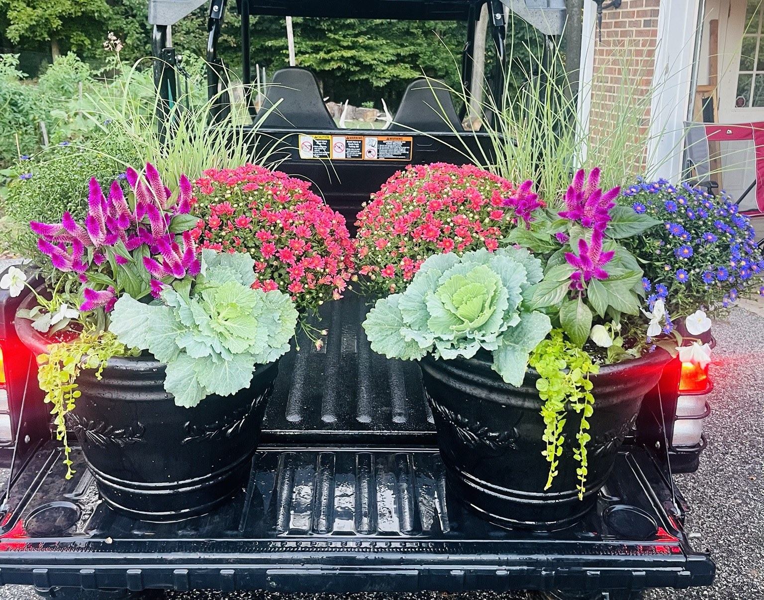 Two black containers filled with fall flowers, ornamental kale, and trailing foliage, ready for delivery