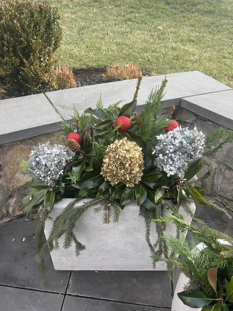 Close-up of a holiday arrangement with evergreen branches, pale blooms, and red berries beside a stone wall