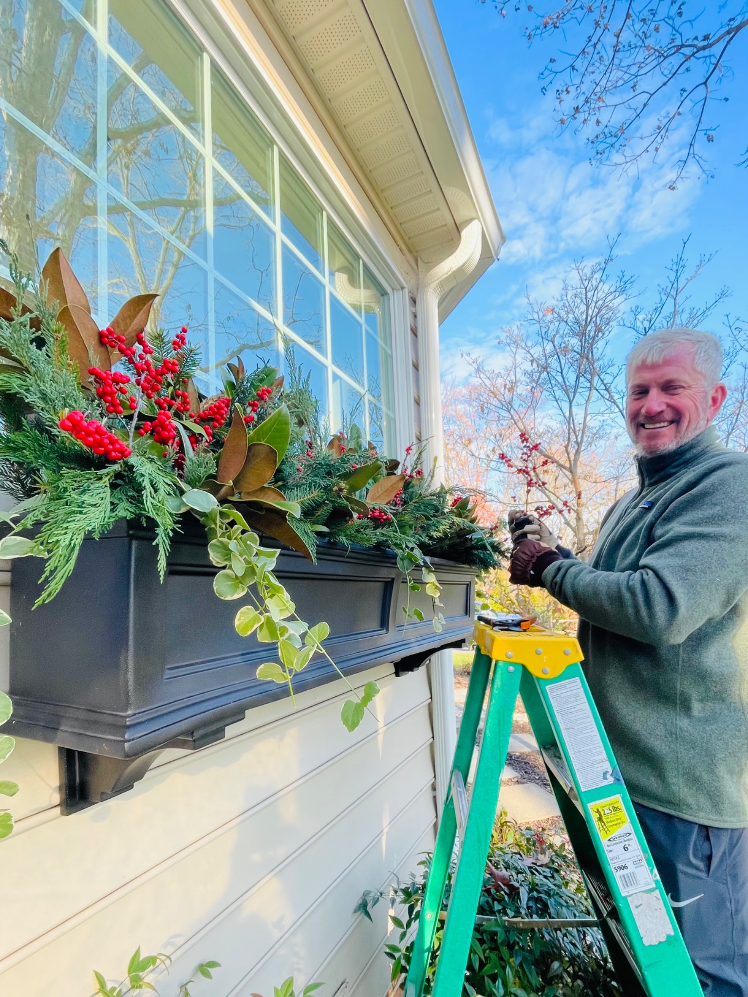 Derek adding finishing touches to a holiday window box filled with evergreens, red berries, magnolia leaves, and trailing foliage