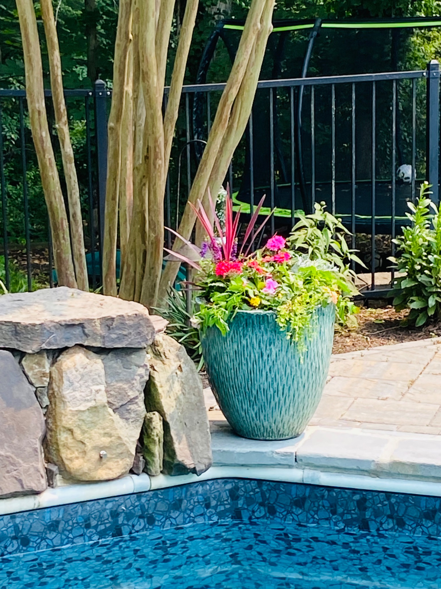 Wider view of a turquoise summer planter with bright blooms beside the pool and stone wall